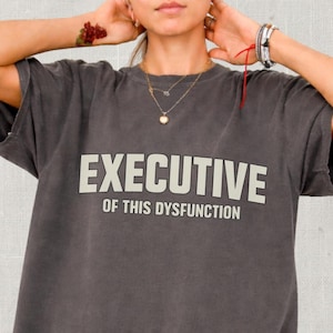May include: Dark gray t-shirt with the words "EXECUTIVE OF THIS DYSFUNCTION" printed in white. The shirt has rolled-up sleeves and a relaxed fit. A person is wearing the shirt.