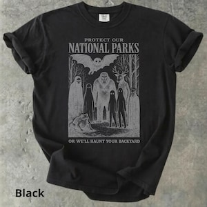 May include: Black t-shirt with a gray graphic featuring cryptids and the text "PROTECT OUR NATIONAL PARKS OR WE'LL HAUNT YOUR BACKYARD." The design includes a bat, Bigfoot, deer, and other mysterious figures.