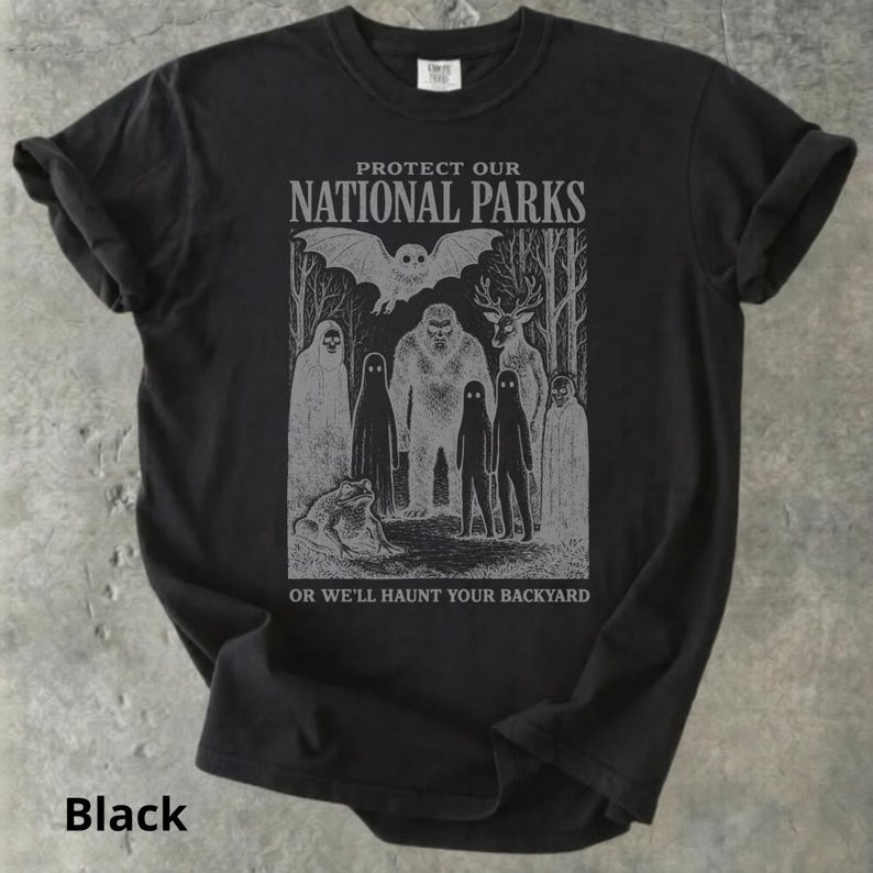 May include: Black t-shirt with a gray graphic featuring cryptids and the text "PROTECT OUR NATIONAL PARKS OR WE'LL HAUNT YOUR BACKYARD." The design includes a bat, Bigfoot, deer, and other mysterious figures.