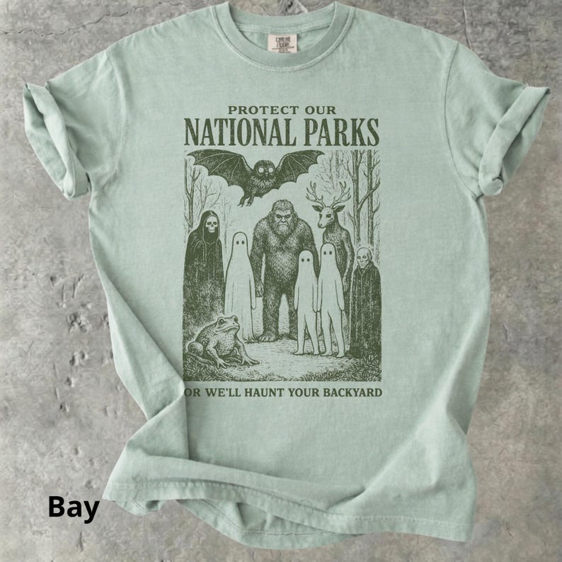 May include: A light green t-shirt with a graphic that reads "PROTECT OUR NATIONAL PARKS" and "OR WE'LL HAUNT YOUR BACKYARD". The graphic features a bat, Bigfoot, a deer, a frog, and several ghost-like figures. The shirt is a solid color.