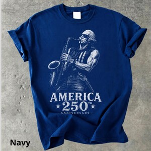 May include: Navy blue t-shirt featuring a white illustration of a person playing a saxophone, with an American flag design on the vest. The text "AMERICA 250th ANNIVERSARY" is printed below the illustration.