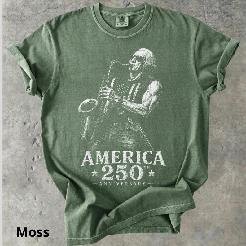 May include: Moss green t-shirt featuring a white graphic of a man playing a saxophone with an American flag design on his vest. The text "AMERICA 250th ANNIVERSARY" is printed below the image. The shirt is made of a soft material.