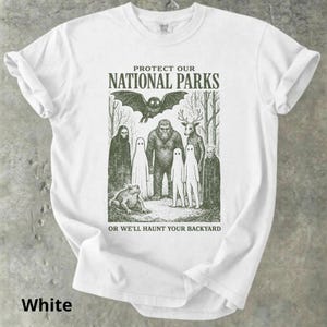 May include: White t-shirt with a green graphic featuring a bat, Bigfoot, deer, ghosts, and a frog. The text reads "PROTECT OUR NATIONAL PARKS" above the image and "OR WE'LL HAUNT YOUR BACKYARD" below. The word "White" is at the bottom.