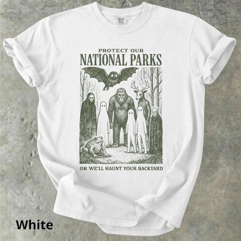 May include: White t-shirt with a green graphic featuring a bat, Bigfoot, deer, ghosts, and a frog. The text reads "PROTECT OUR NATIONAL PARKS" above the image and "OR WE'LL HAUNT YOUR BACKYARD" below. The word "White" is at the bottom.