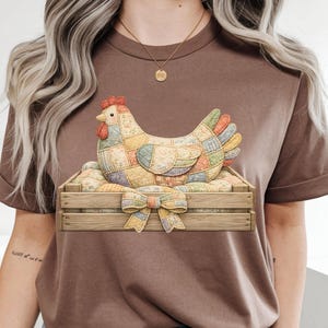 May include: Brown t-shirt featuring a colorful patchwork hen sitting in a wooden crate with a bow. The hen has a red comb and a variety of patterned patches. The crate is filled with eggs.