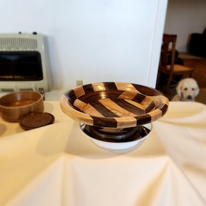 Puede incluir: Un cuenco de madera con un patrón a rayas en tonos marrones y madera clara. El cuenco está sobre un pedestal blanco y está sentado sobre un mantel blanco.