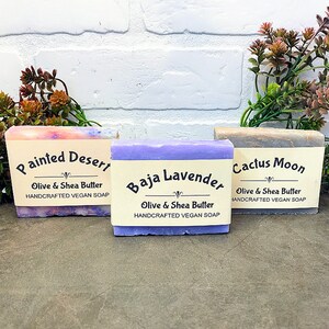 May include: Three bars of handmade vegan soap with labels that read "Painted Desert", "Baja Lavender", and "Cactus Moon". Each bar is made with olive and shea butter.