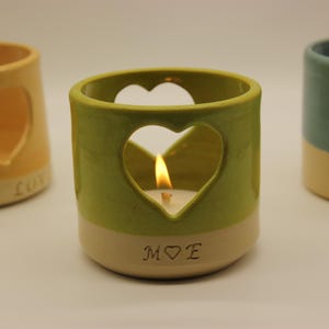 Peut inclure: Trois bougeoirs en céramique avec des découpes en forme de cœur. Le bougeoir du centre est vert et blanc avec une bougie allumée à l'intérieur et l'inscription "M♥E". Le bougeoir de gauche est jaune et blanc avec l'inscription "MY LOVE". Le bougeoir de droite est bleu et blanc avec l'inscription "MOM".