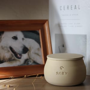 Peut inclure: Une urne en céramique beige mat avec un couvercle, gravée du nom ROXY et d'une petite icône de patte. À côté se trouve un cadre photo en bois contenant la photographie d'un chien blanc aux yeux sombres et au nez noir, près d'un livre blanc.