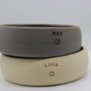 Op de afbeelding: Twee keramische huisdierkommen, een grijs en een crèmekleurig, met poot afdrukken en de namen "MAX" en "LUNA" gegraveerd.