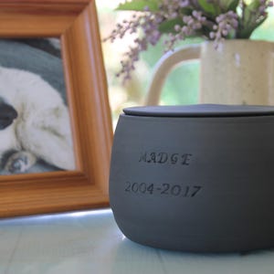 Könnte beinhalten: Dunkelgraue Urne für Haustiere mit Deckel, beschriftet mit dem Namen "MADGE" und den Jahren "2004-2017". Im Hintergrund ist ein gerahmtes Foto eines weißen Hundes sowie eine Blumenvase zu sehen.