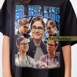 May include: Black t-shirt featuring a graphic of a person wearing glasses, with the text "DR. MEL KING" in blue. The design includes multiple images of the person and lightning bolt effects, creating a vintage style.