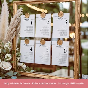 May include: A gold-framed mirror seating chart featuring six white rectangular cards labelled Table 1 through 6. Each card lists names in black text and is secured at the top with a round gold wax seal. The display is decorated with pampas grass.
