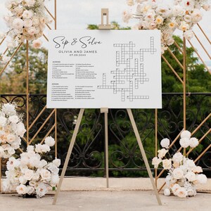 Large sip and solve wedding crossword puzzle displayed on an easel among white floral wedding arrangements. Perfect as a custom puzzle game sign for wedding receptions or bridal showers.