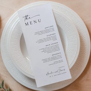 May include: A white menu card with black text listing the appetizers, starters, main course, and desserts for a wedding reception. The menu includes items like toasted bread with mozzarella, a hearty soup, steak, wedding cake, and chocolate chip cookies. The bottom of the card reads "Amelia and James 08-15-2024 Miami, Florida."