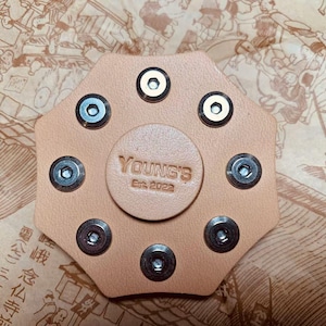 May include: A tan leather fidget spinner with eight silver metal accents. The spinner has the text "YOUNG'S Est. 2022" in the center.