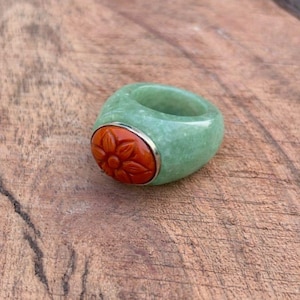Puede incluir: Un anillo de jade verde con un diseño de flor naranja tallada engastada en un bisel plateado. El anillo se exhibe sobre una superficie de madera. El anillo es una pieza de joyería única.