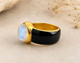 Puede incluir: Un anillo de tono dorado con una banda negra lisa y pulida, que presenta una piedra lunar grande, redonda y tallada en cabujón. La piedra lunar muestra un aspecto blanco translúcido con un brillo azul iridiscente, engastada en oro.