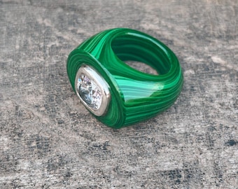 May include: A vibrant green malachite ring with a square, clear gemstone set in a silver bezel. The ring's band features a swirling pattern of light and dark green, creating a striking visual effect. This unique piece of jewelry is a statement accessory.