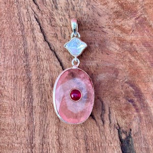 Puede incluir: Colgante de plata con una gran piedra ovalada rosa facetada, una pequeña piedra redonda roja y una piedra cuadrada iridiscente. El colgante cuelga de una fianza de plata. Las piedras están engastadas en plata. El colgante está sobre una superficie de madera.