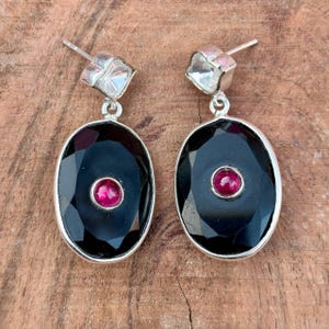 Puede incluir: Un par de pendientes de plata con cierre de poste. Cada pendiente presenta una piedra negra ovalada facetada con una pequeña gema roja redonda en el centro. Los pendientes se muestran sobre una superficie de madera.
