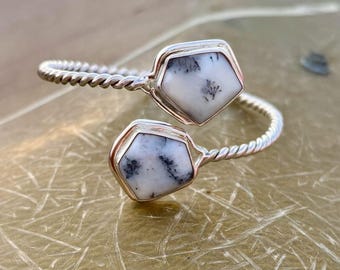 Peut inclure: Un bracelet manchette argenté orné de deux pierres blanches en forme de pentagone avec des inclusions noires. Les pierres sont serties dans des chatons argentés et reliées par une bande torsadée en argent. Le bracelet est conçu pour s'enrouler autour du poignet.