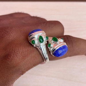 May include: Two silver rings with blue and green gemstones. Each ring features a large, oval blue stone and several smaller green stones. The rings are displayed on a dark skin tone.