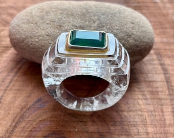 May include: A clear, carved ring with a rectangular, emerald-green gemstone set in a gold-colored bezel. The ring is on a wooden surface, with a smooth, gray stone in the background.