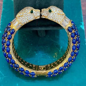 Puede incluir: Una pulsera dorada con dos cabezas de pantera engastadas con cristales transparentes y ojos verdes. La pulsera está adornada con piedras preciosas ovaladas de color azul oscuro. El diseño es intrincado y lujoso, mostrando una mezcla de elegancia y estilo audaz.