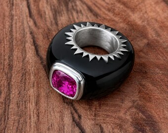 May include: A chunky black ring featuring a smooth, rounded band with a silver-toned sunburst or spiked detail around the inner opening. The ring is set with a single, square-cut, vibrant pink gemstone encased in a polished silver-toned metal frame.