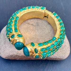 May include: A gold-toned hinged bracelet with emerald-green gemstones. The bracelet features a series of oval-shaped green stones set in a repeating pattern along the outer edges. The bracelet is resting on a gray stone.