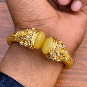 May include: A gold-toned cuff bracelet featuring two large, smooth, oval-shaped yellow stones at the open ends. The metal surface is textured with fine ridges and decorated with small, clear, round inset stones surrounding the yellow centres.