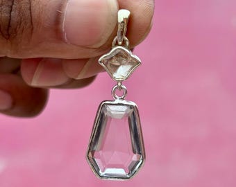Può includere: Un pendente trasparente e sfaccettato con montatura color argento. Il pendente ha un design geometrico con una pietra più piccola a forma di diamante sopra una pietra esagonale allungata più grande. Lo sfondo è rosa pieno.