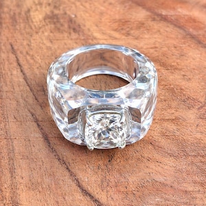 May include: Clear, faceted ring with a large, square-cut centre stone. The ring's band is thick and also faceted, creating a sparkling effect. The ring is set against a wooden surface.