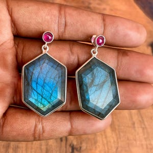 May include: A pair of silver earrings featuring large, faceted, hexagonal labradorite stones with blue and green iridescence. Each earring has a small, round, red gemstone at the top.