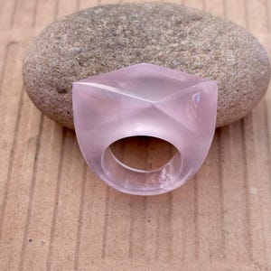 以下が含まれることがあります： 幾何学模様の、半透明の薄いピンク色の指輪。 指輪は正方形の上面と円形の開口部があります。 指輪は、滑らかな灰色の石の隣の茶色の表面に表示されています。
