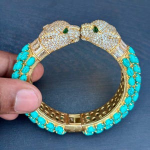 May include: Gold-toned bracelet featuring two leopard heads encrusted with clear crystals and green eyes. The bracelet is adorned with turquoise-colored stones and has a hinged clasp. A luxurious piece of jewelry.