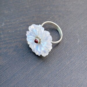 May include: A silver ring with a carved moonstone flower design, with a small red gemstone at the centre. The moonstone has an iridescent quality, and the ring is set on a dark grey surface.
