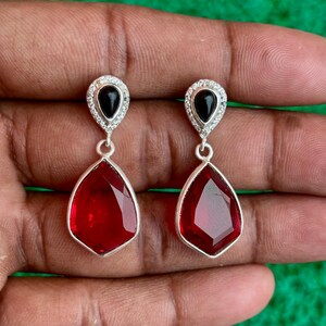 Puede incluir: Un par de pendientes colgantes plateados. Cada pendiente presenta una piedra preciosa roja oscura facetada en forma de lágrima, suspendida debajo de una piedra negra más pequeña engastada en un halo de cristales transparentes. Los pendientes se muestran sobre un fondo verde.