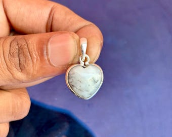 May include: A heart-shaped pendant with a silver-colored bail. The pendant features a white stone with gray inclusions, set in a silver bezel. The pendant is held against a purple background.