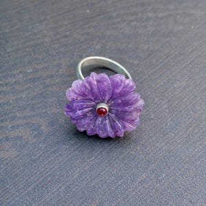 Könnte beinhalten: Ein silberner Ring mit einem geschnitzten lila Blumenmuster und einem kleinen roten Edelstein in der Mitte. Die Blume ist aufwendig detailliert und schafft ein einzigartiges Schmuckstück. Der Ring ist vor einem dunkelgrauen Hintergrund platziert.