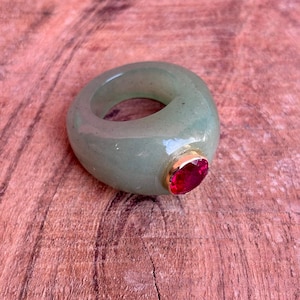 Puede incluir: Un anillo verde claro con un engaste dorado y una gran gema roja de forma ovalada. El anillo está hecho de un material translúcido y descansa sobre una superficie de madera. La gema es el punto focal del anillo.