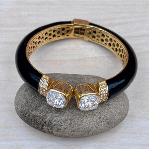 Puede incluir: Un brazalete rígido articulado en tono dorado con un acabado de esmalte negro liso, un interior con patrón de panal y dos terminales abiertos, cada uno con una gran piedra transparente de corte cuadrado flanqueada por cristales pavé.