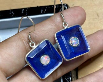 May include: A pair of square earrings with a deep blue gemstone and a central opal. The earrings have a silver setting and a hook closure. The opal has a mix of colors. The earrings are a jewelry item.
