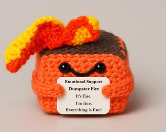 May include: A handmade orange crochet figure with a brown top and black eyes, holding a sign that reads "Emotional Support Dumpster Fire." The figure has orange and yellow flames on top. The sign also says "It's fine. I'm fine. Everything is fine!"