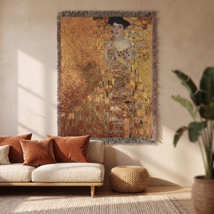 Peut inclure: Une tapisserie murale tissée représentant une reproduction du Portrait d'Adele Bloch-Bauer I de Gustav Klimt. Le textile rectangulaire affiche une silhouette dans une robe dorée aux motifs géométriques complexes, avec une bordure à franges.