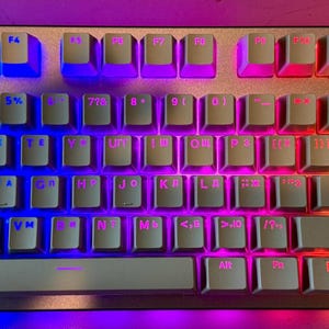 May include: A white mechanical keyboard with rainbow colored backlighting. The keyboard has a standard layout with function keys, number keys, and arrow keys. The keys are white with black lettering.