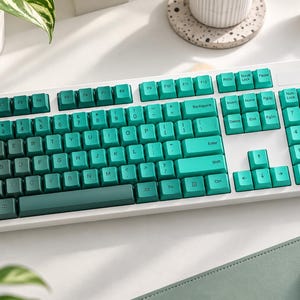 May include: A full-sized mechanical computer keyboard with a white frame and a gradient of teal and dark green keycaps. The layout includes a standard QWERTY arrangement, a numeric keypad on the right, and function keys across the top row.