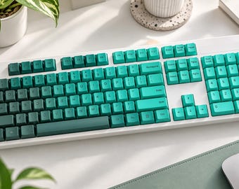 May include: A full-sized mechanical computer keyboard with a white frame and a gradient of teal and dark green keycaps. The layout includes a standard QWERTY arrangement, a numeric keypad on the right, and function keys across the top row.