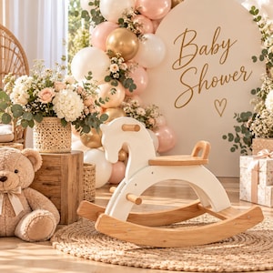 May include: A white and natural wood rocking horse with a smooth finish, featuring a curved seat, two horizontal handles, and arched rockers. The horse is positioned on a round woven rug in a room decorated with balloons and a Baby Shower sign.